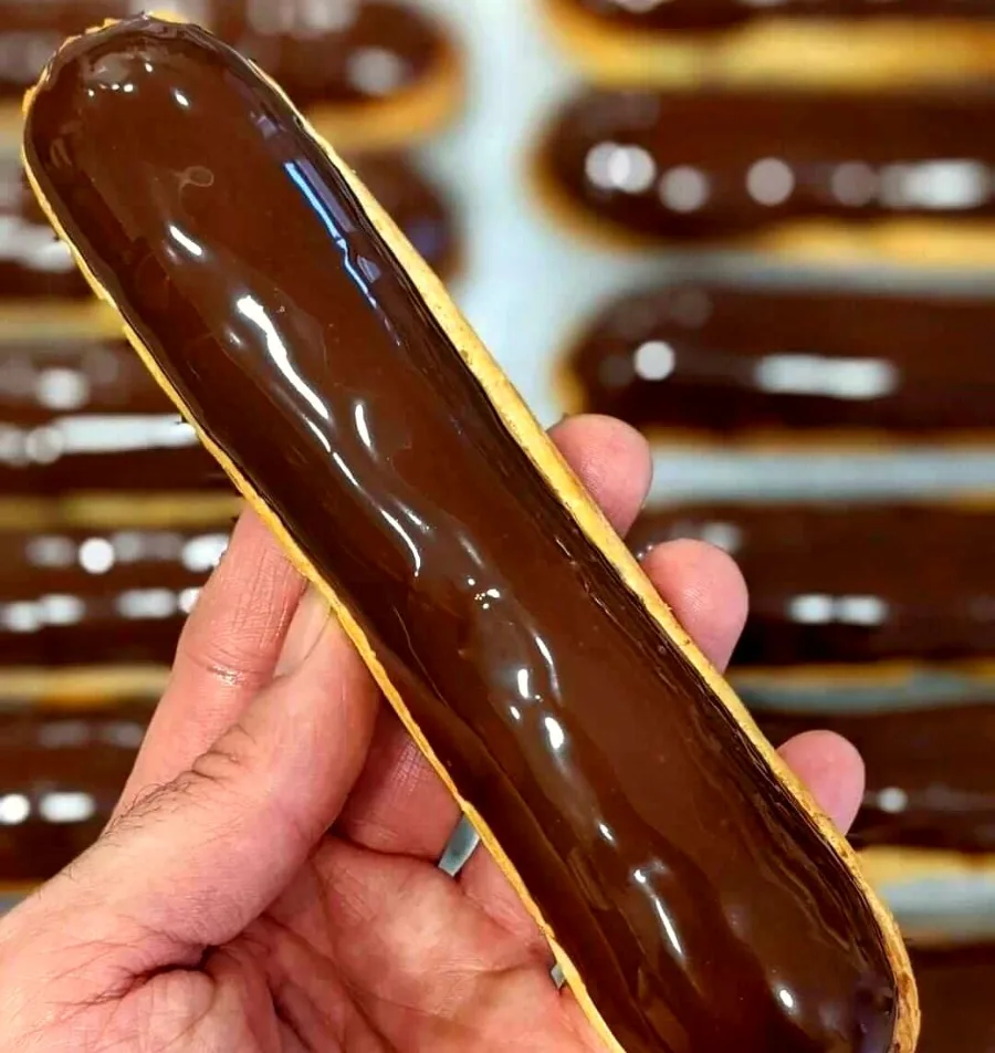 Éclair glacé au chocolat tenu à la main, devant une plaque d'éclairs